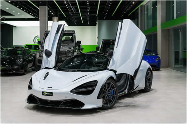 White McLaren 720S with dihedral doors open, displayed in a luxury Dubai car showroom with G-Class and Porsche in the background